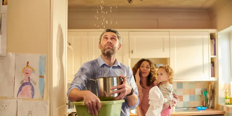 El hombre que recoge agua en una olla y cubo de un techo de fuga, con una mujer y un niño preocupados en una cocina, lo que ilustra el impacto del daño de la lluvia y la necesidad de asistencia de seguros en Houston.